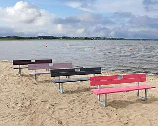 Vier farbige Bänke stehen am Ufer eines Gewässers.